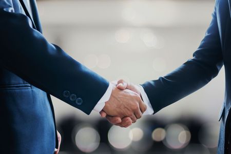 Two businesspeople in dark suits shaking hands in a bright office setting