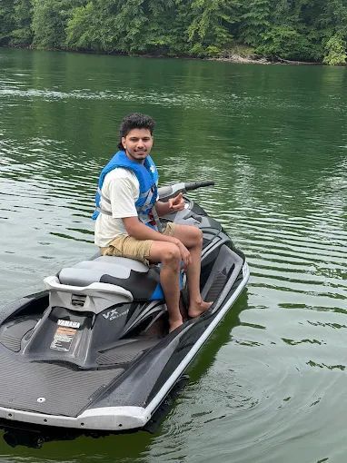 Person sitting on a black jet ski on calm green water near a forested shore.