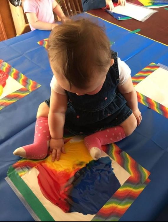 Baby sitting on a table, exploring colorful paint with hands, wearing a denim dress and pink socks.