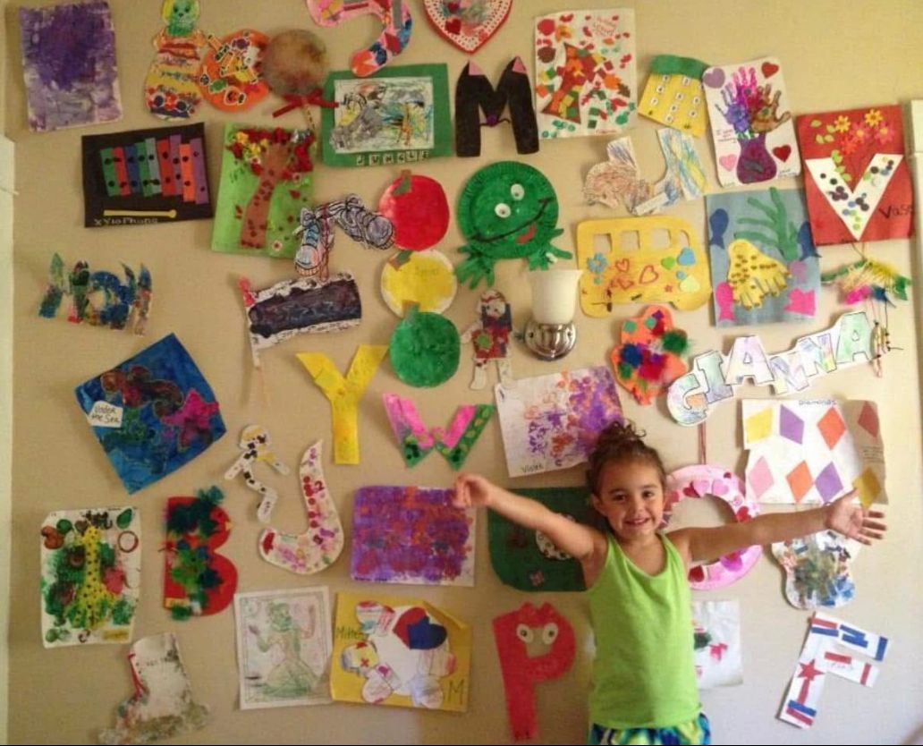 Young girl smiles with arms wide, standing before a wall covered in colorful children's artwork.