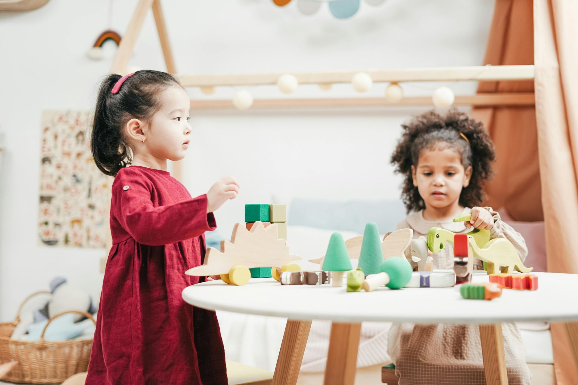 Two young girls playing with wooden toys on a round white table in a playroom.