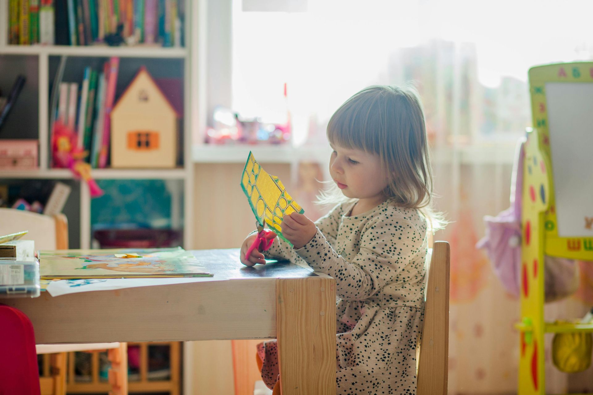 Young child sitting at a table, cutting paper with scissors. Light streams in from a window.