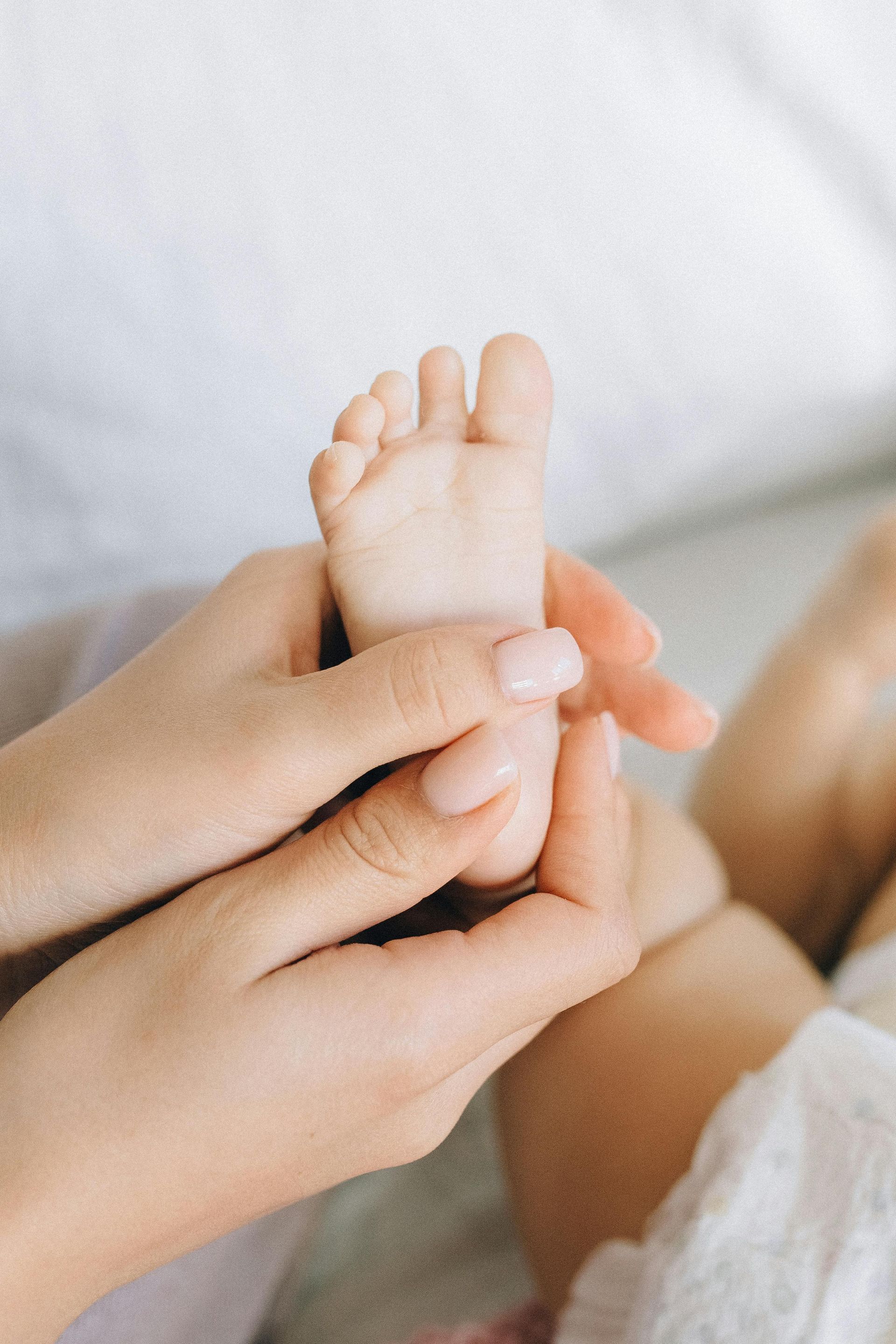 Hands holding a baby's tiny foot, pale skin. White background, soft focus.