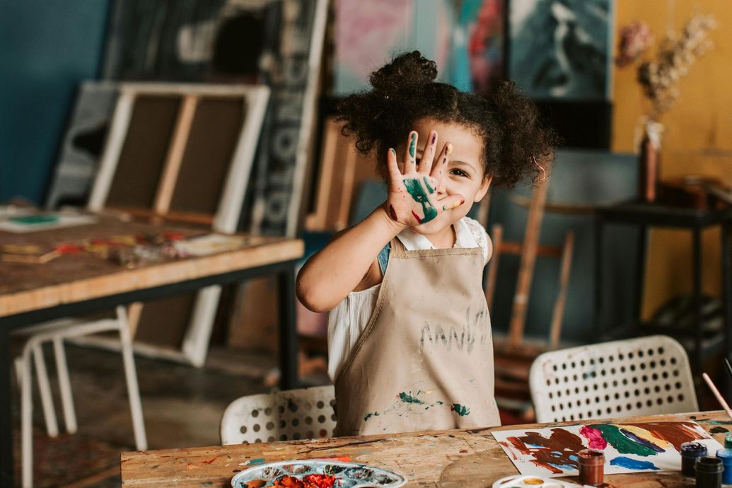 Young child in art smock smiles, holds up paint-covered hand; art studio setting.