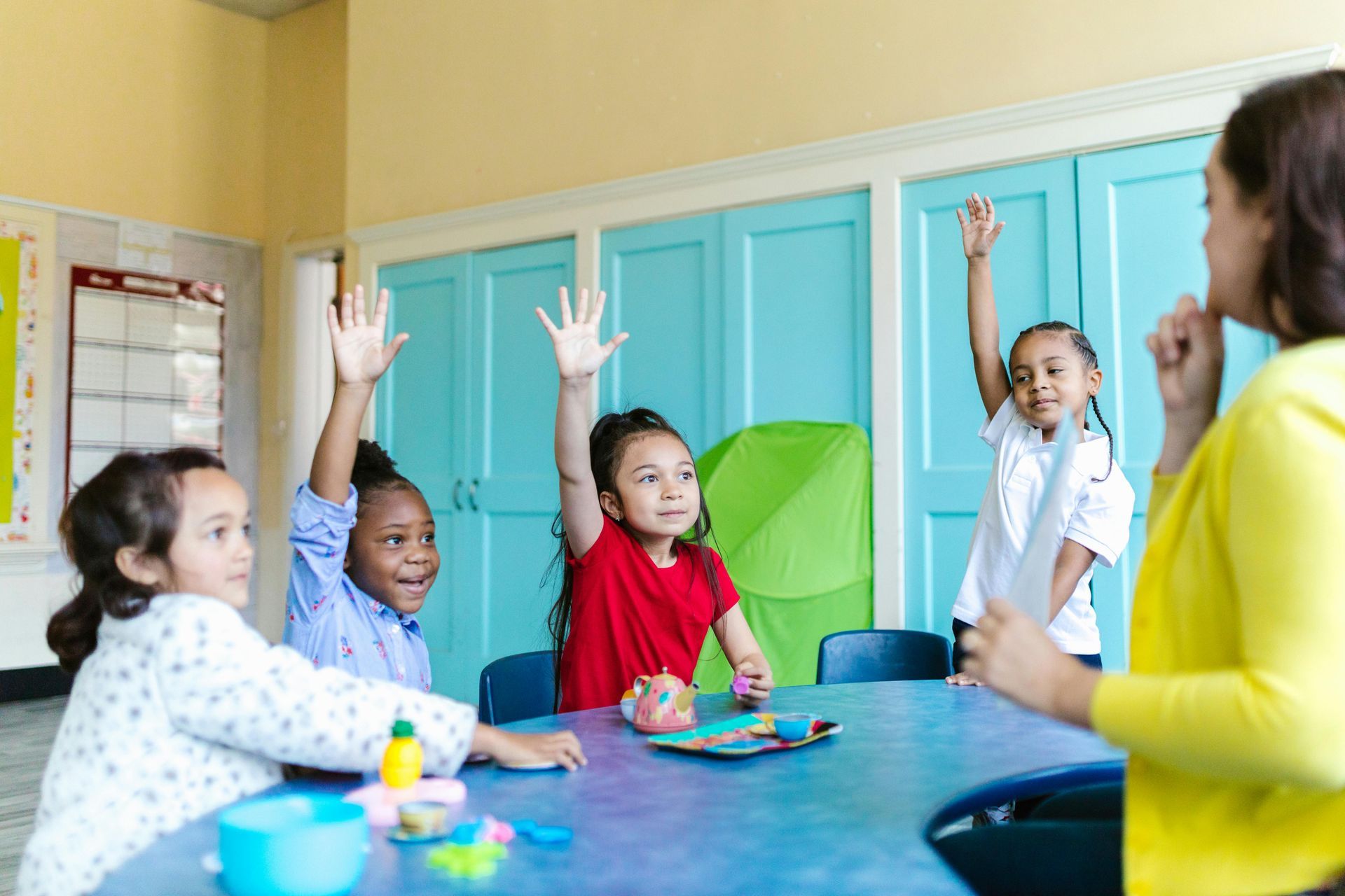 Children raising hands in classroom, teacher with yellow sweater, blue table.