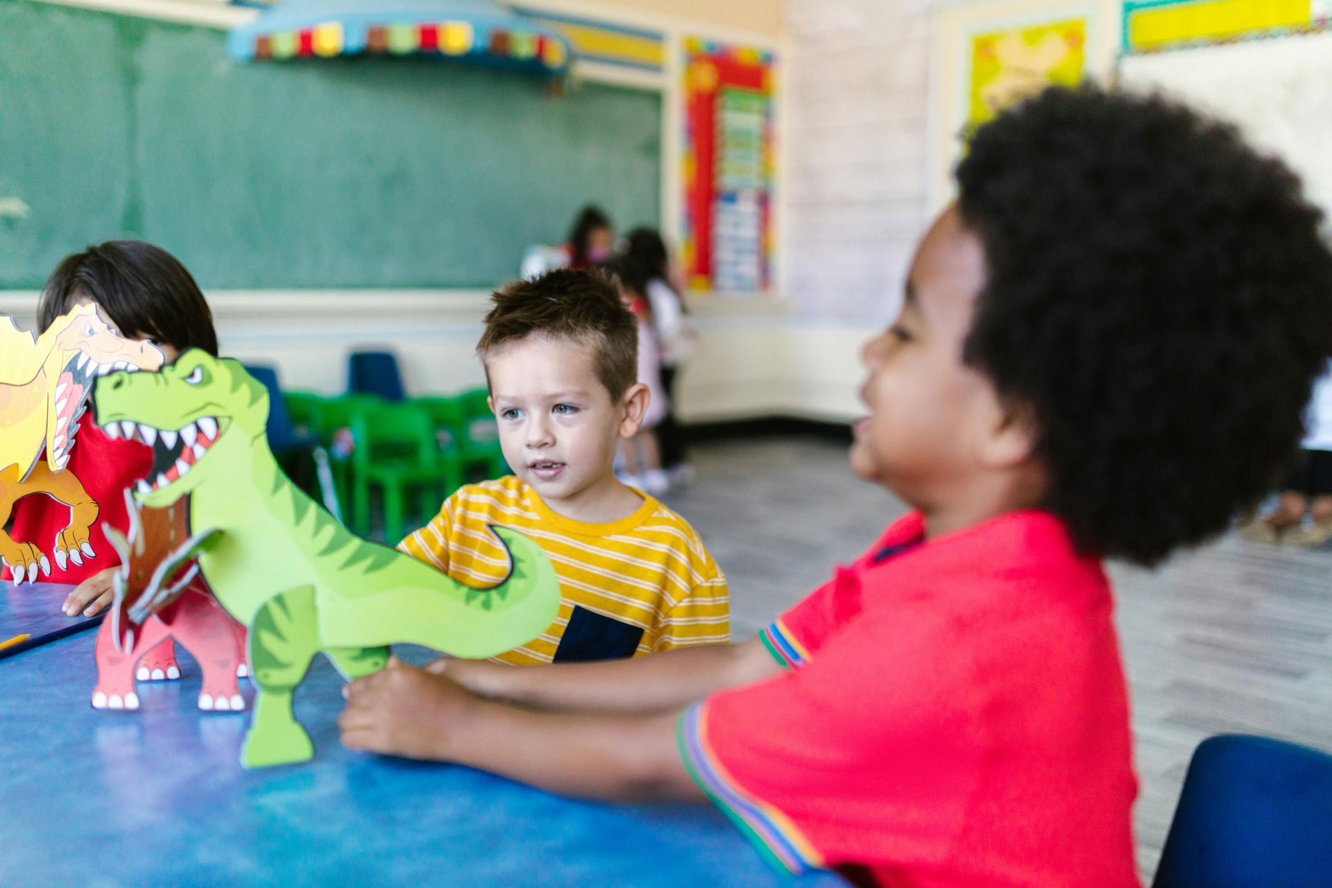 Children in classroom playing with green dinosaur cutouts at a table.