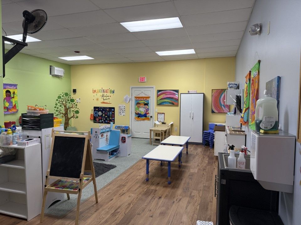 Inside a brightly colored preschool classroom: tables, art, toys, and a water dispenser are visible.