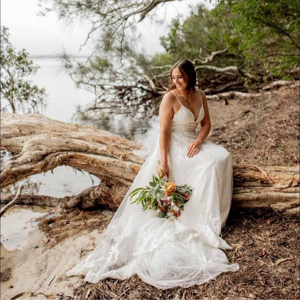 A bride in a wedding dress is sitting on a log by the water holding a bouquet of flowers. — Vanity Skin and Beauty Forster in Forster, NSW
