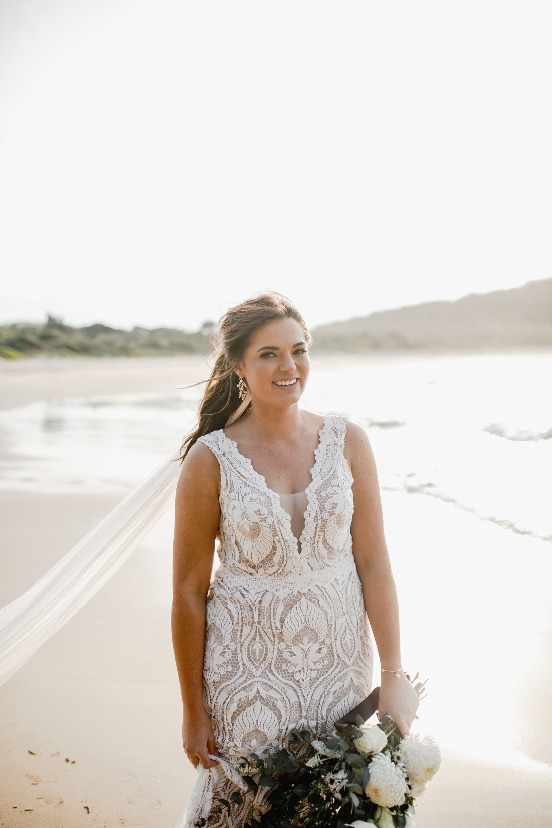 A bride in a wedding dress is standing on the beach holding a bouquet of flowers. — Vanity Skin and Beauty Forster in Forster, NSW