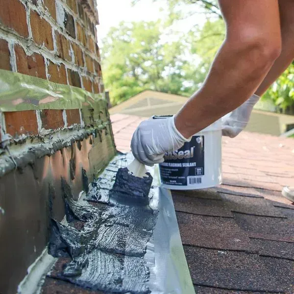 Roofer applies sealant to roof flashing near a brick chimney, wearing a hard hat and safety vest.
