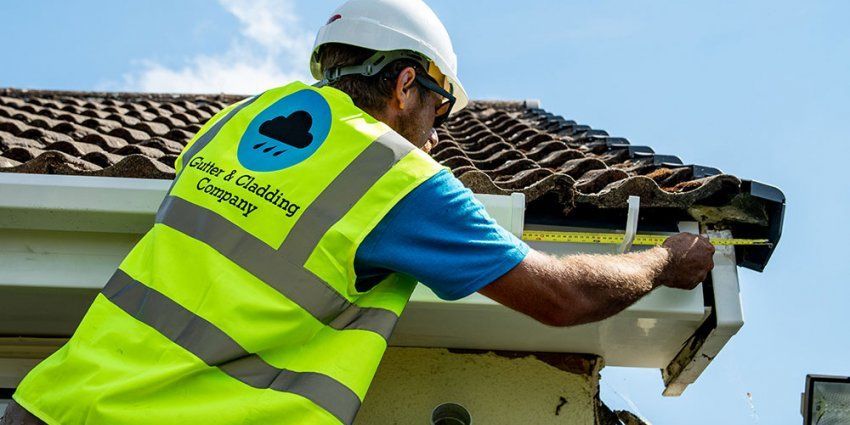 Construction worker on a ladder, caulking a gutter on a house. Blue sky, white vest, and hard hat.