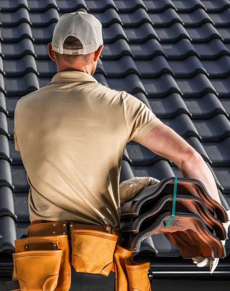 Roofer in safety gear installing shingles on a house roof at sunset, suburban neighborhood background.