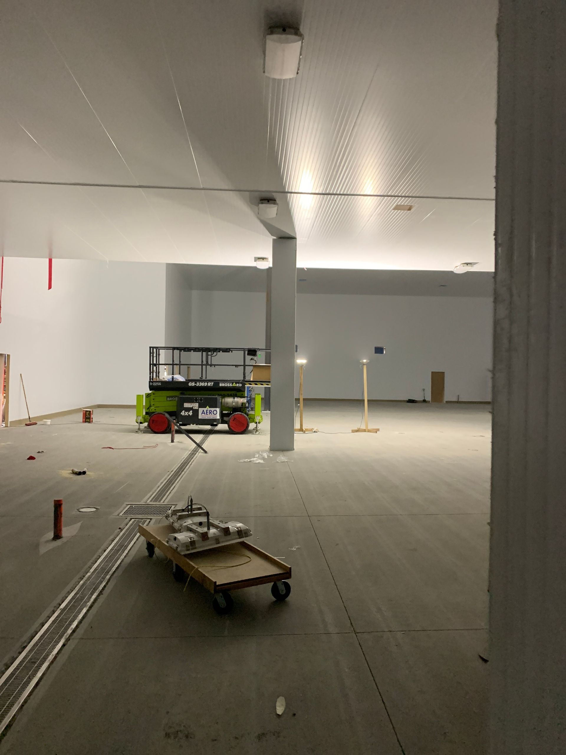 An unfinished indoor construction site with a scissor lift, a wooden dolly with tools, and exposed concrete flooring.