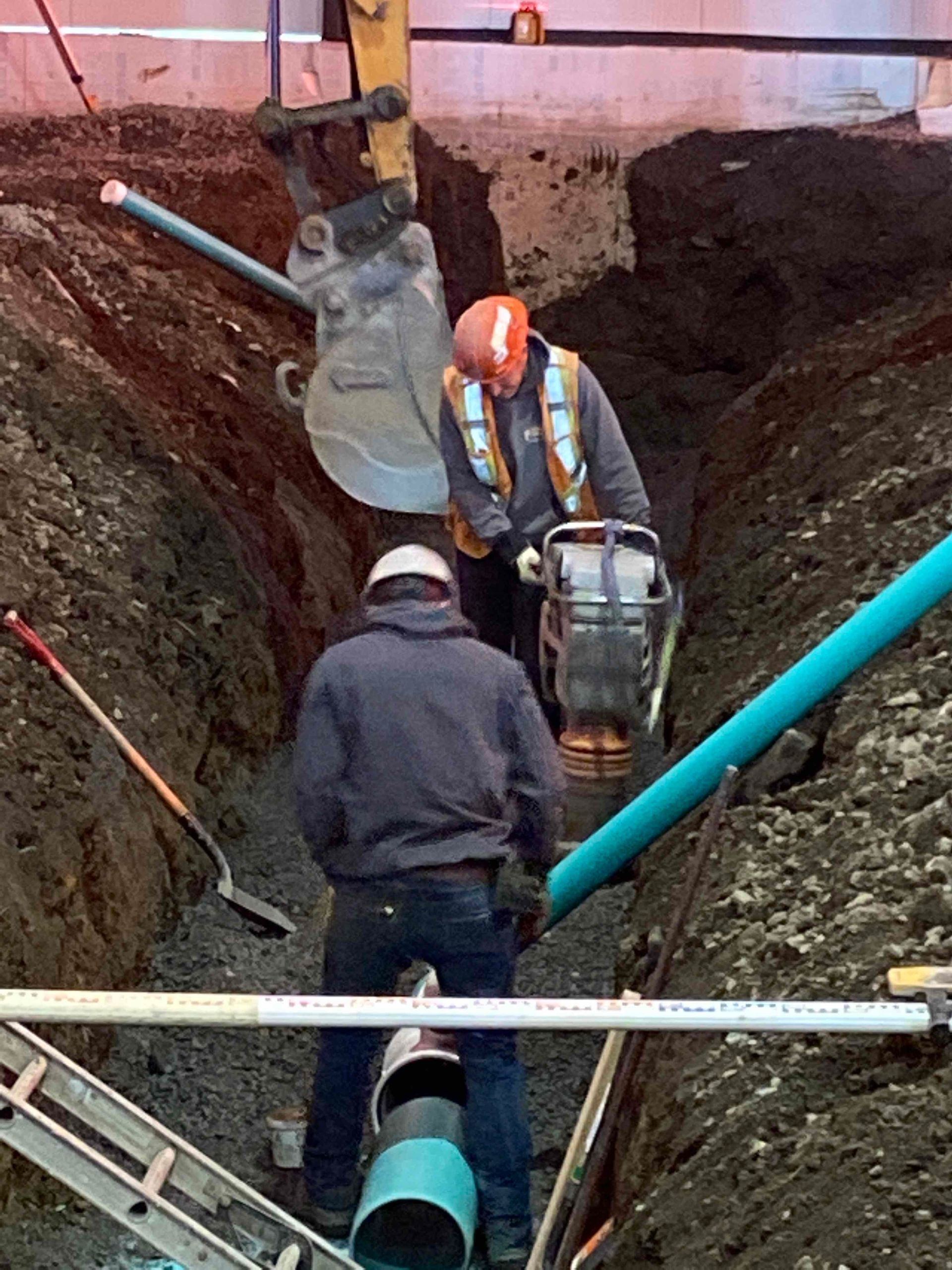 Des ouvriers des services publics installent des tuyaux bleus dans une tranchée profonde, à l'aide d'échelles et d'engins de terrassement à proximité.