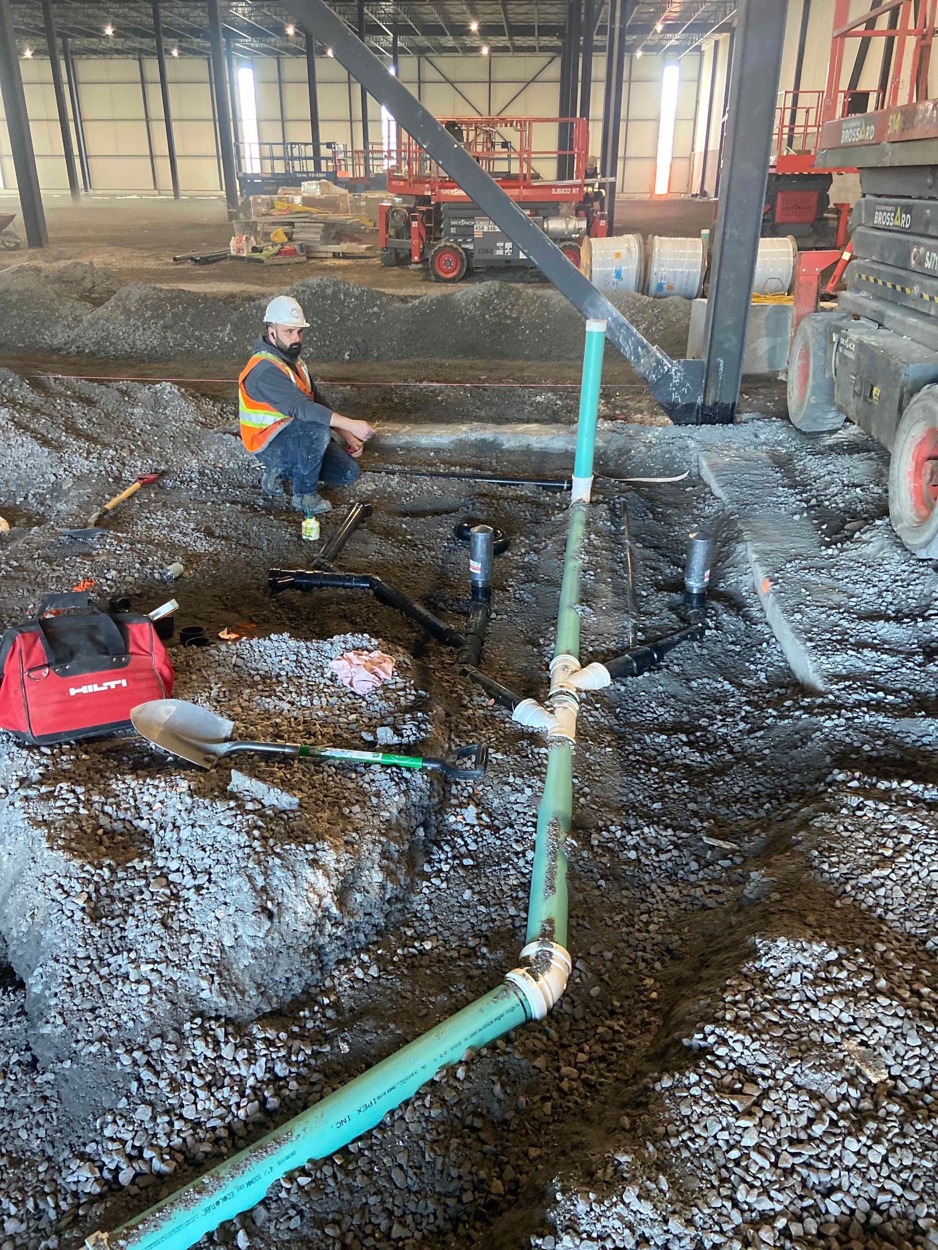 Des ouvriers casqués inspectent un chantier boueux où circulent des tuyaux et des engins de terrassement.