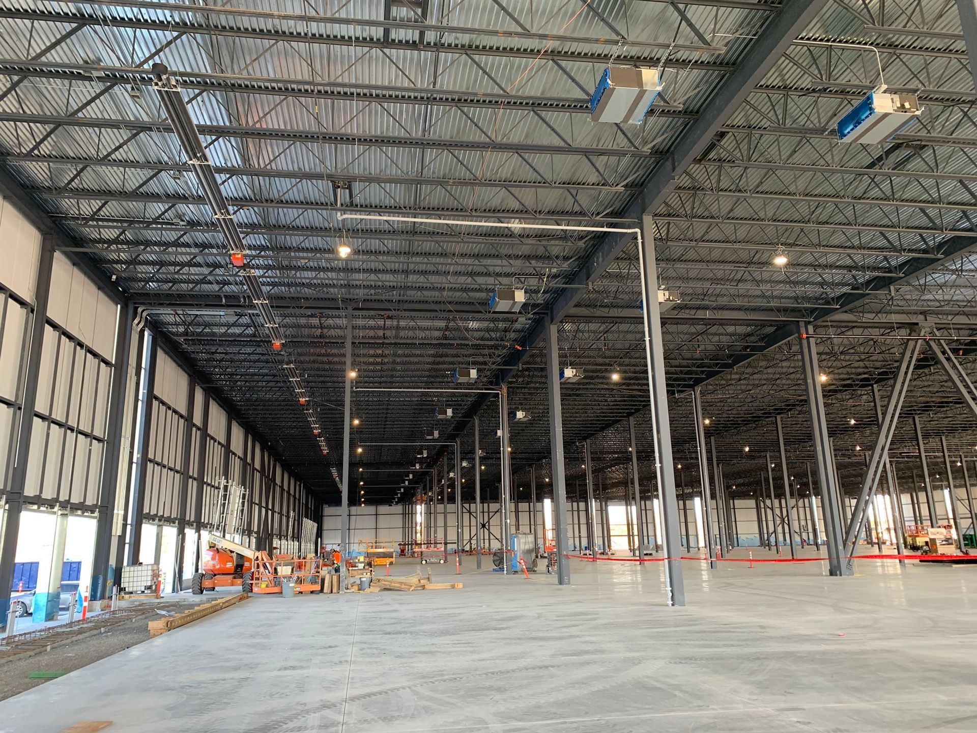 Large, unfinished interior of a warehouse space with high, exposed steel truss ceilings, concrete floors, and support beams.