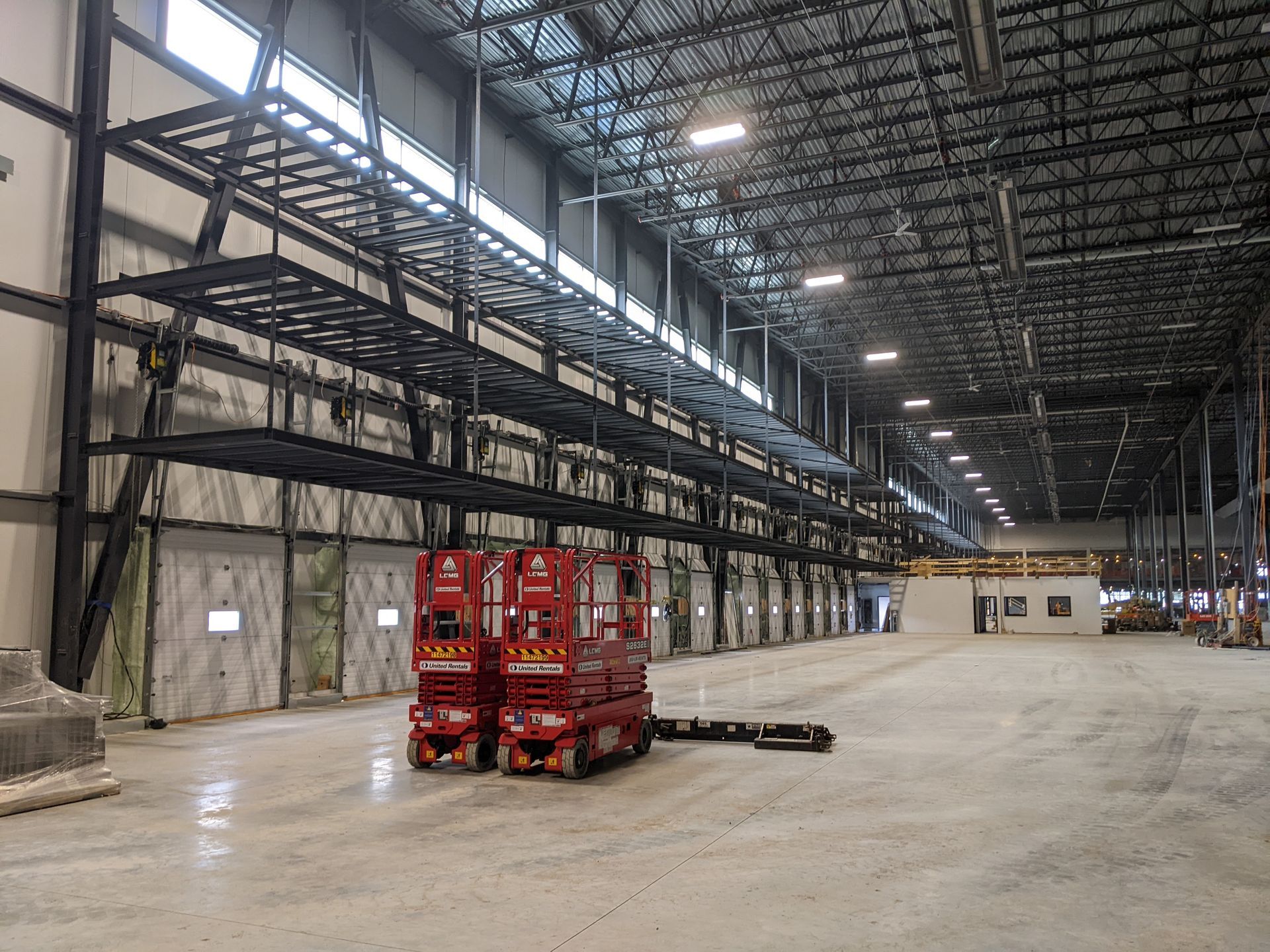 Two red scissor lifts sit on a concrete floor in a large, unfinished warehouse with metal racking along the wall.