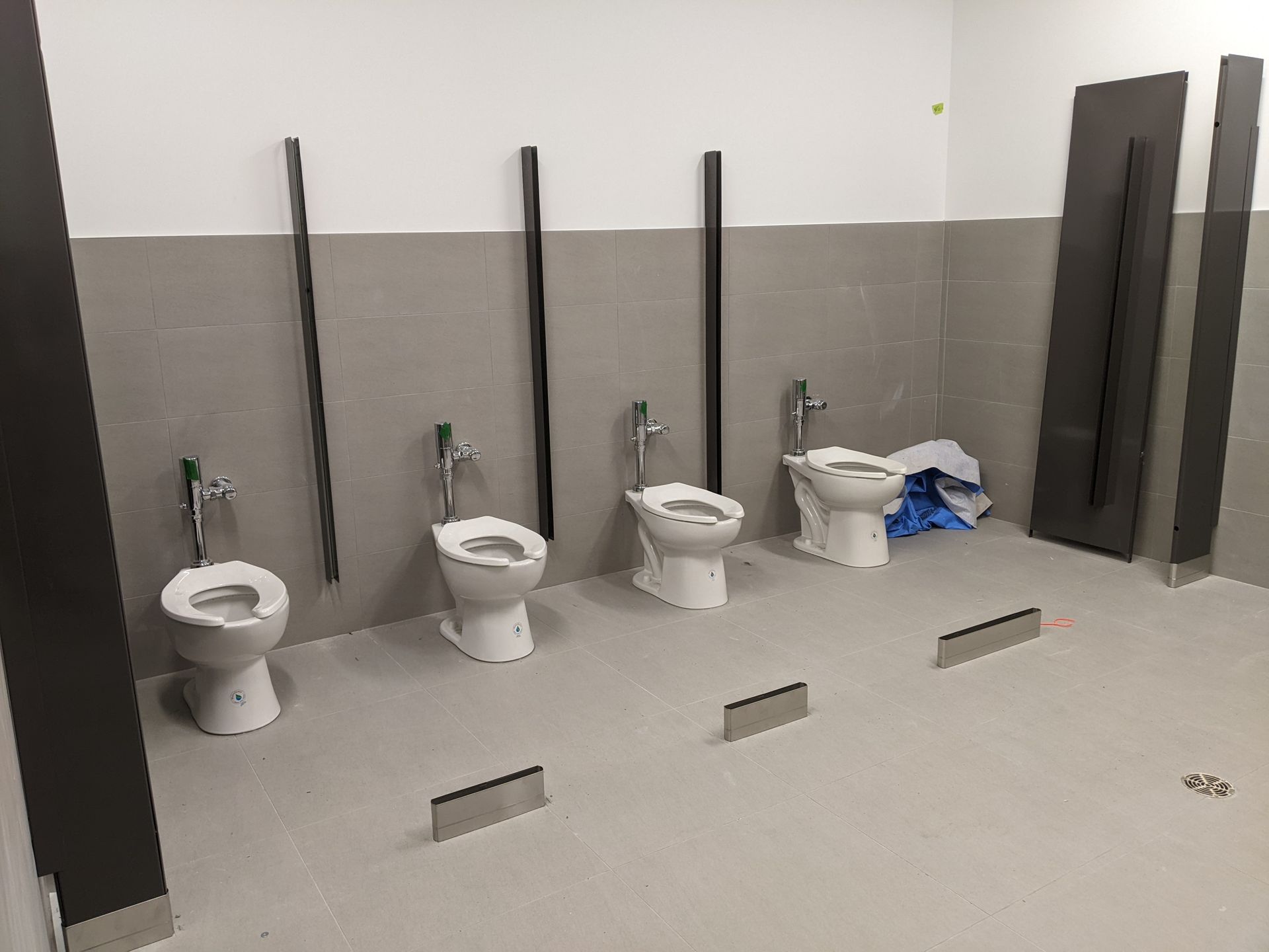 Toilettes publiques en construction, avec quatre toilettes blanches alignées contre un mur carrelé gris.