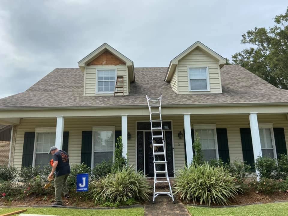Person on ladder, roof repair on beige house with columns and shutters. Overcast sky.