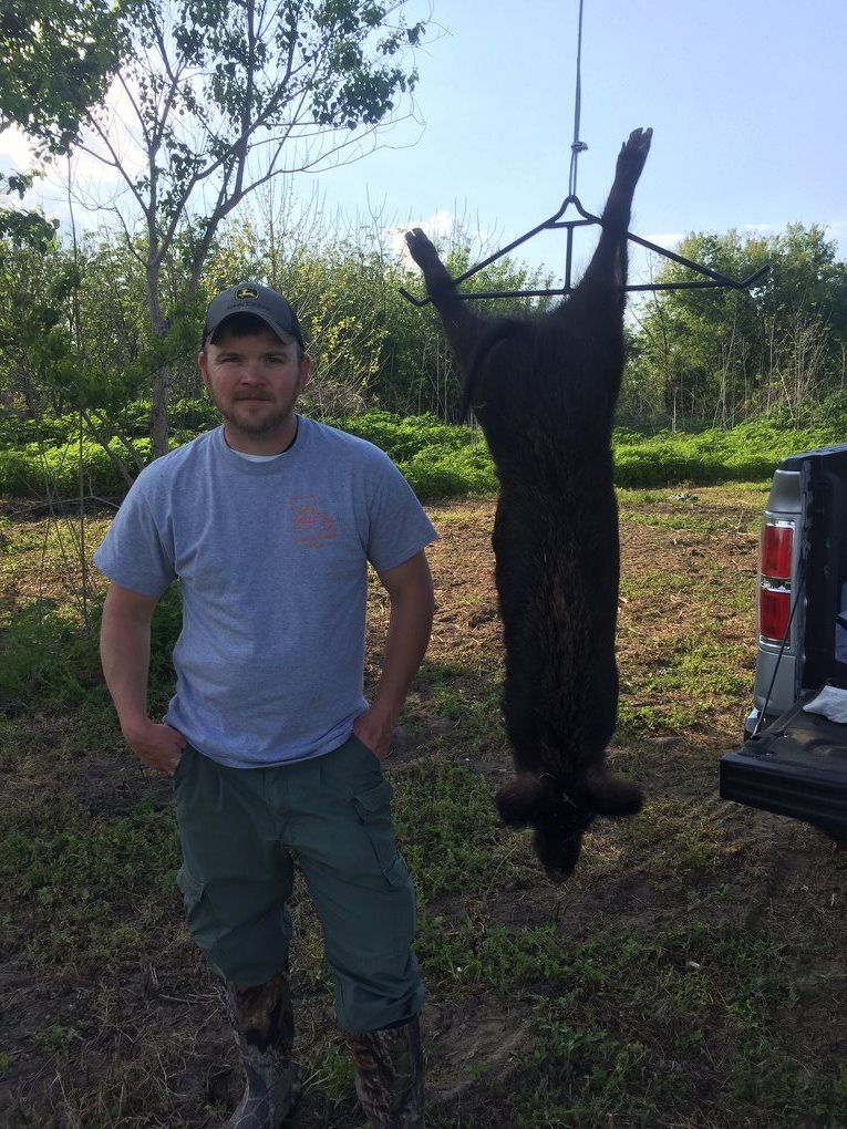 Man poses beside a large, hanging, dark-furred hog near a truck and trees.