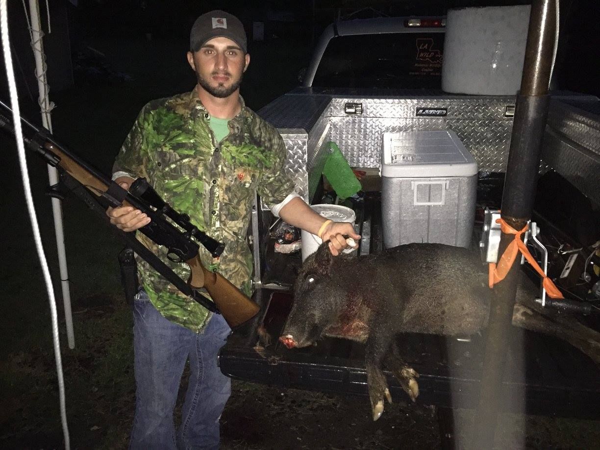 Man with rifle and dead wild pig in truck bed. He is wearing camouflage, a cap, and smiling.