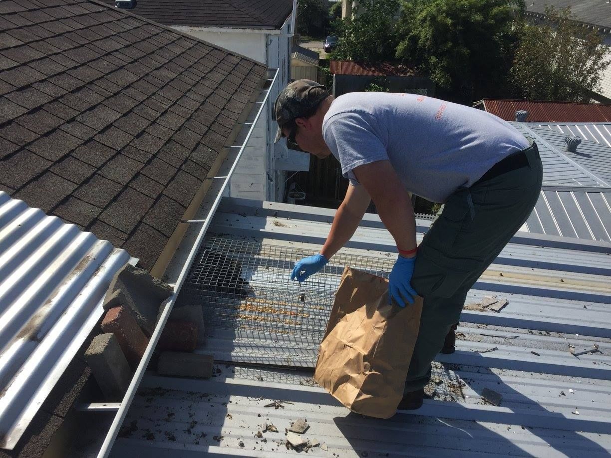 Man on a rooftop cleaning debris from a metal roof; wearing gloves, sunglasses, and a light colored shirt.