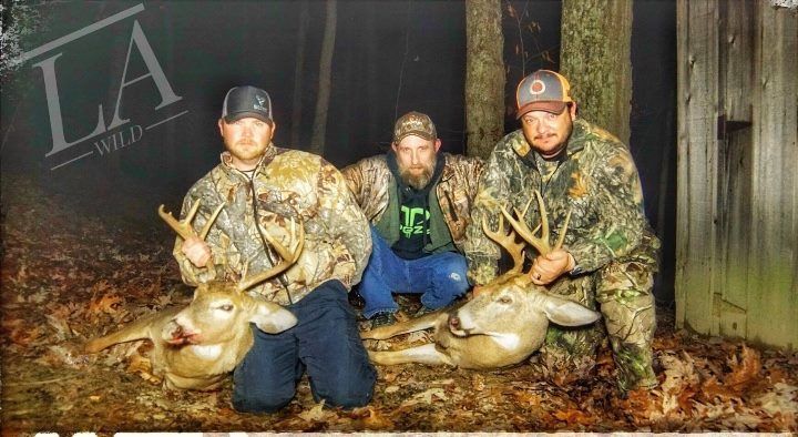 Three men in camo with deer they hunted, in a forest, at night.