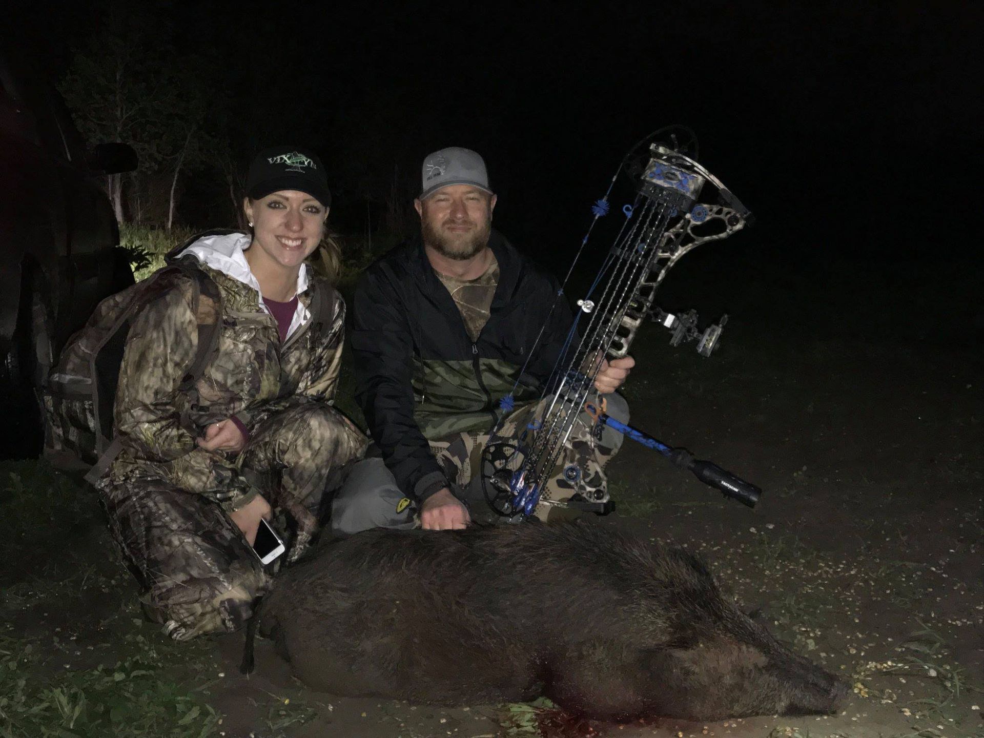 A man and woman in camo kneel with a wild boar at night, man holding a bow.