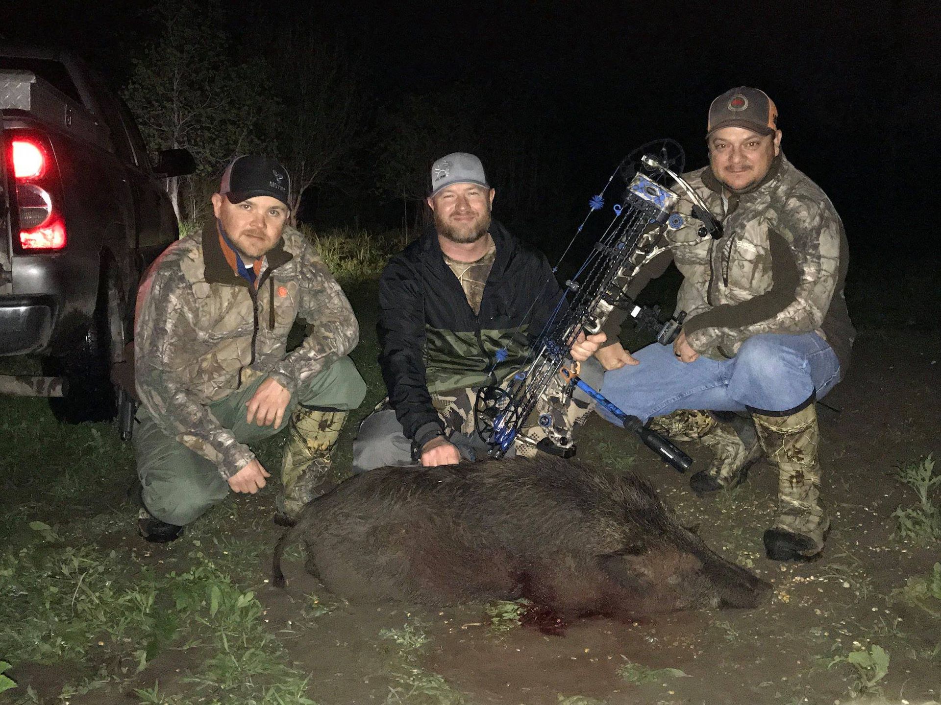 Three men pose with a harvested wild boar and a bow. They are in camouflage gear at night near a truck.