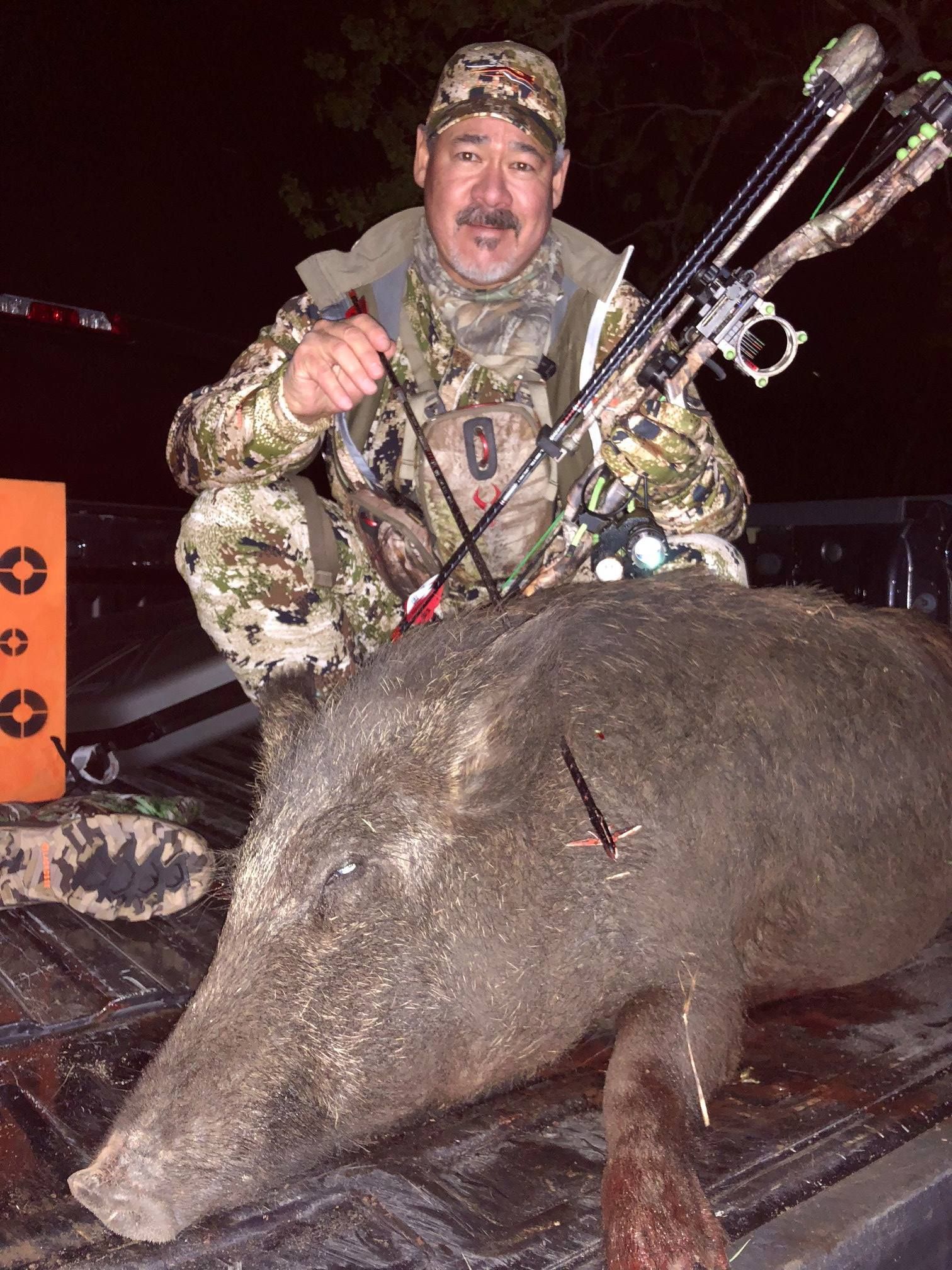 Hunter in camo with a bow, kneeling next to a large wild hog on a truck bed.
