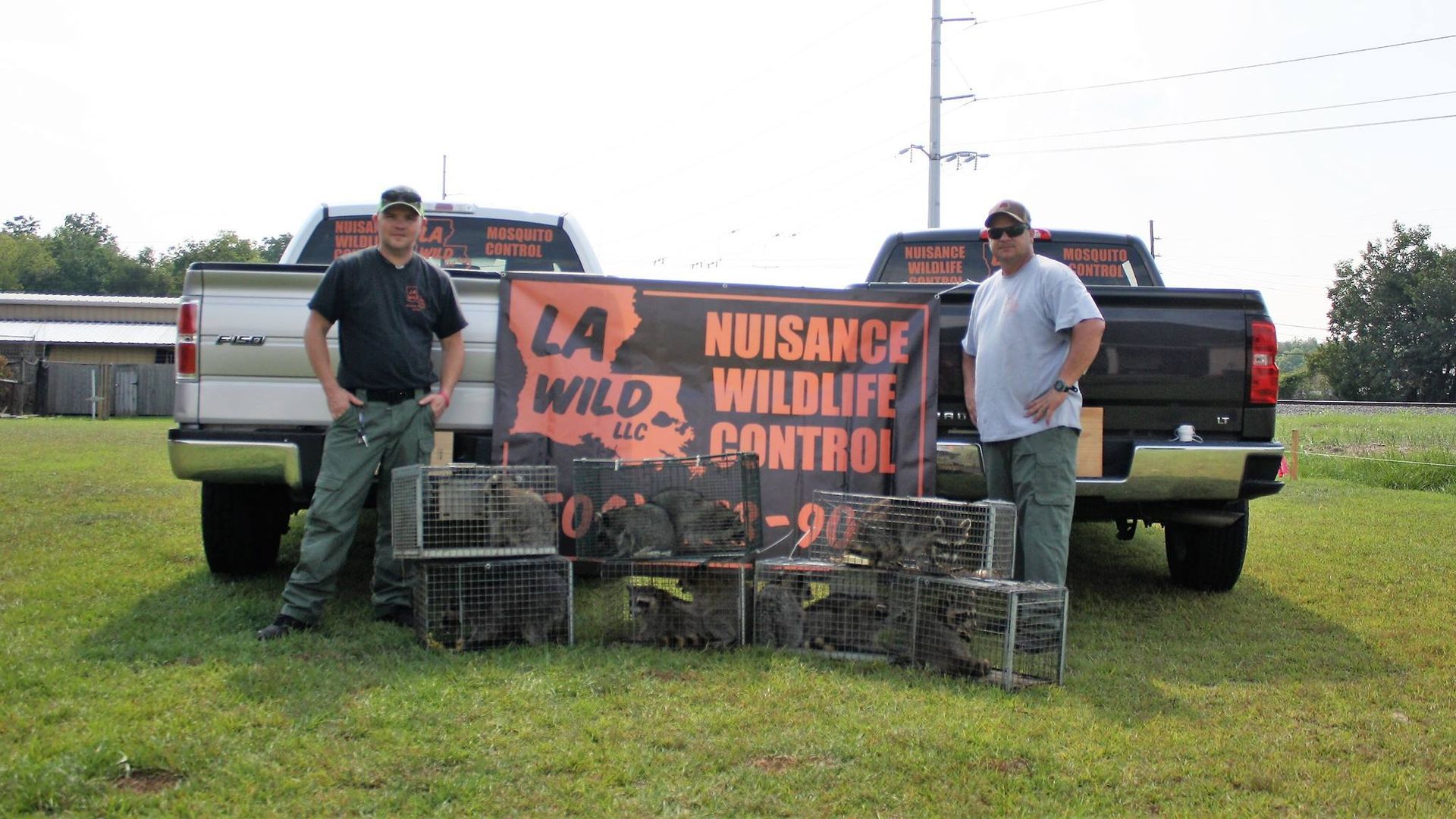 Two men pose with animal traps in front of trucks, a sign for 