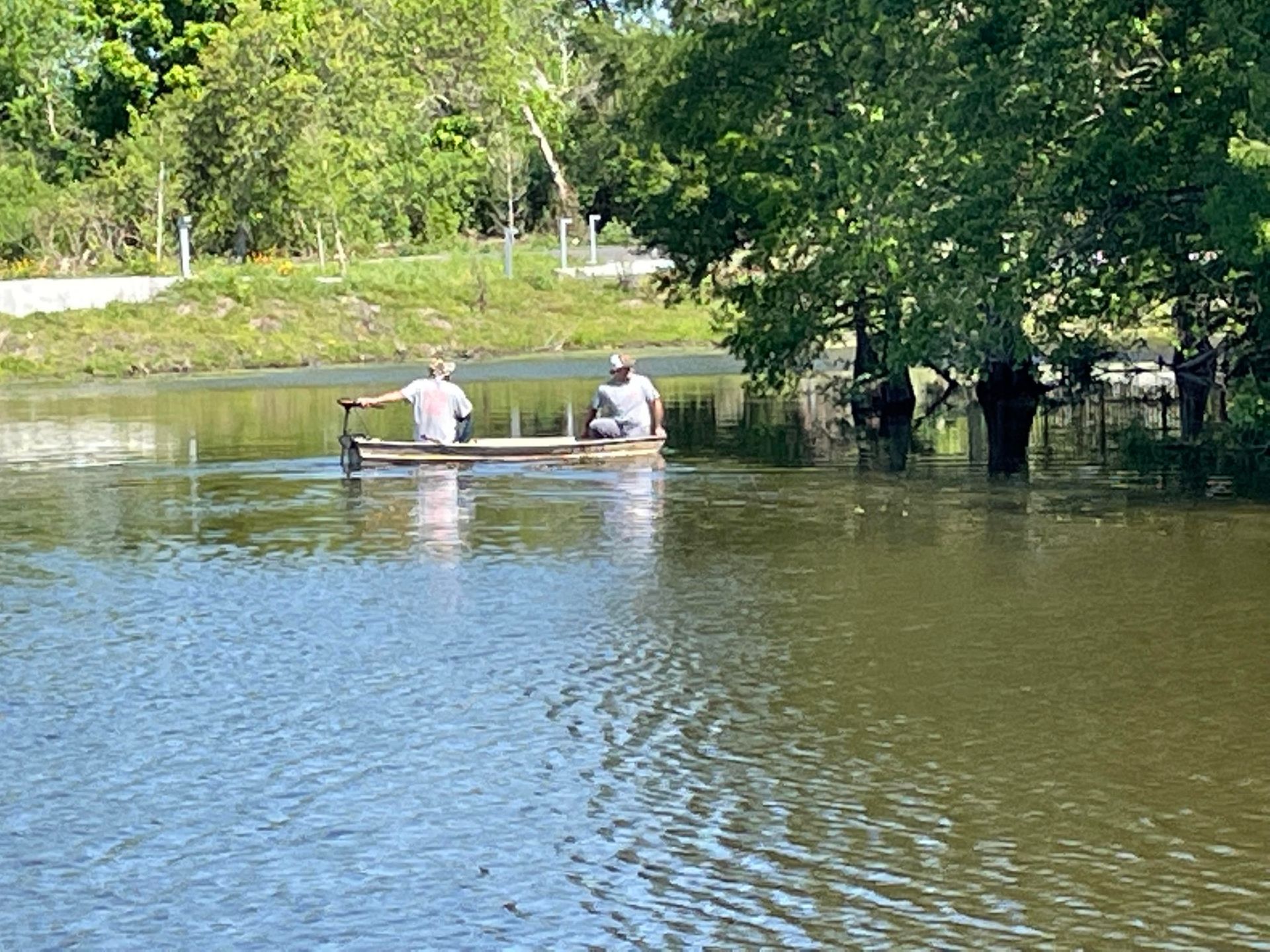 Two people on a raft in a flooded area. Dark water, green trees, and overcast sky.