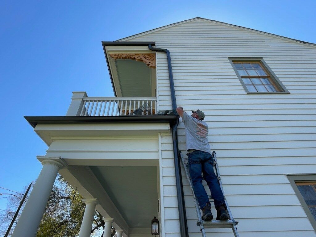 Man on a ladder installing black gutter on a white house with porch and blue sky.
