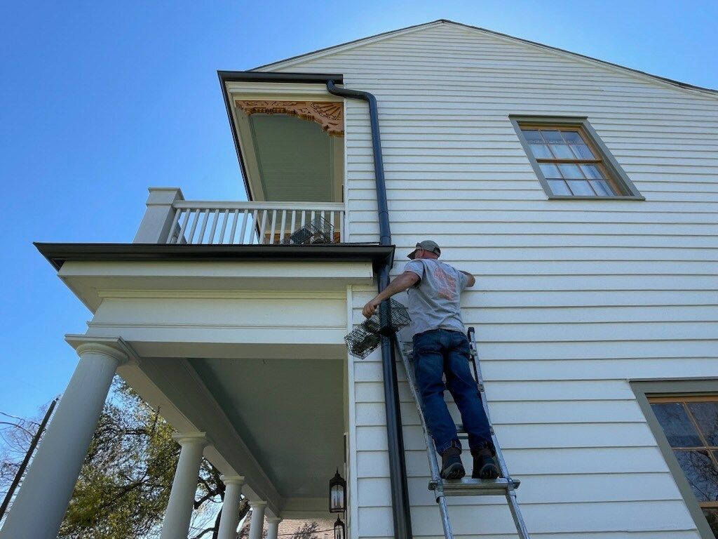 A man installs a black gutter on a white two-story house, using a ladder. Blue sky.