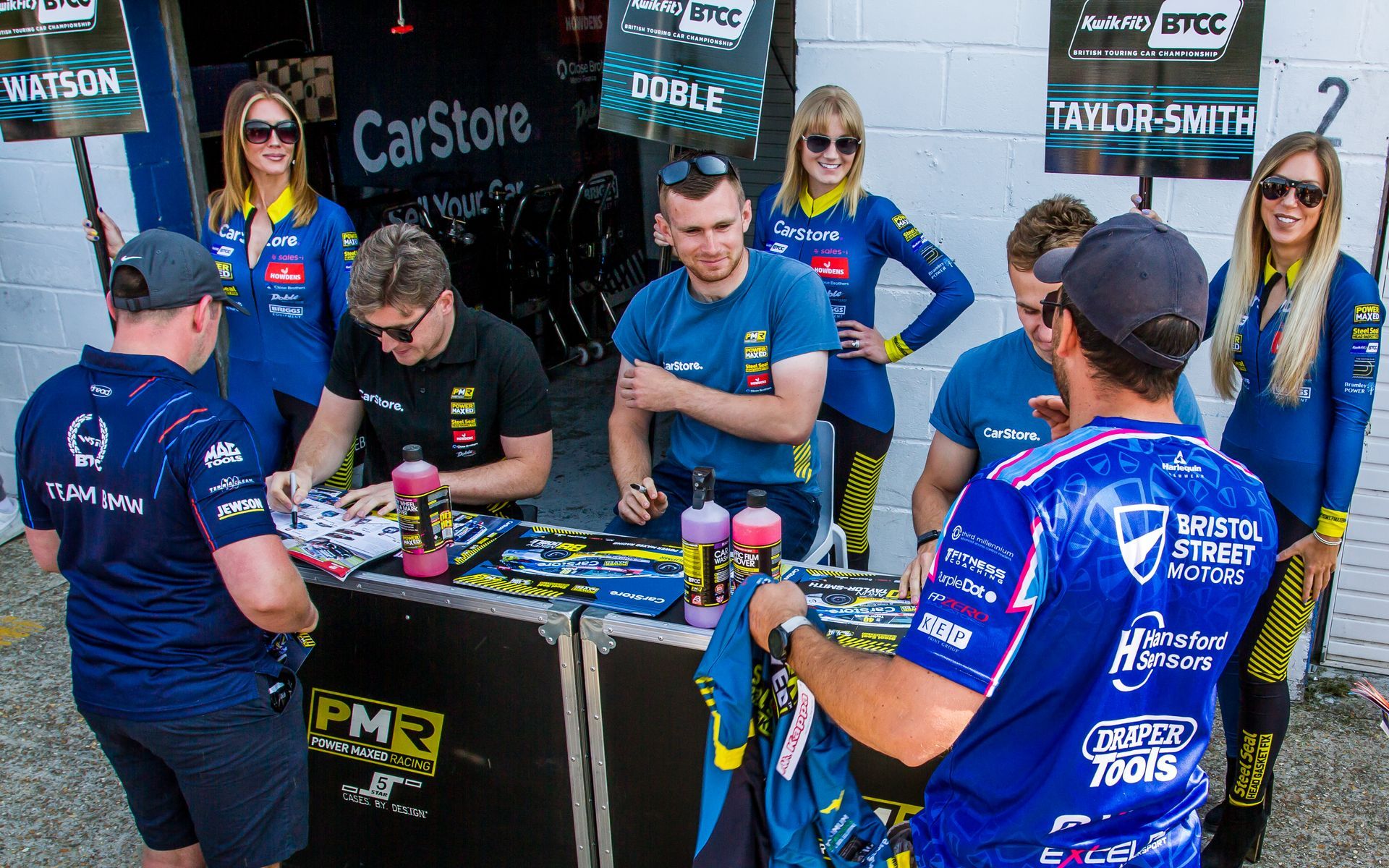 A group of people are standing around a table in front of a car store.