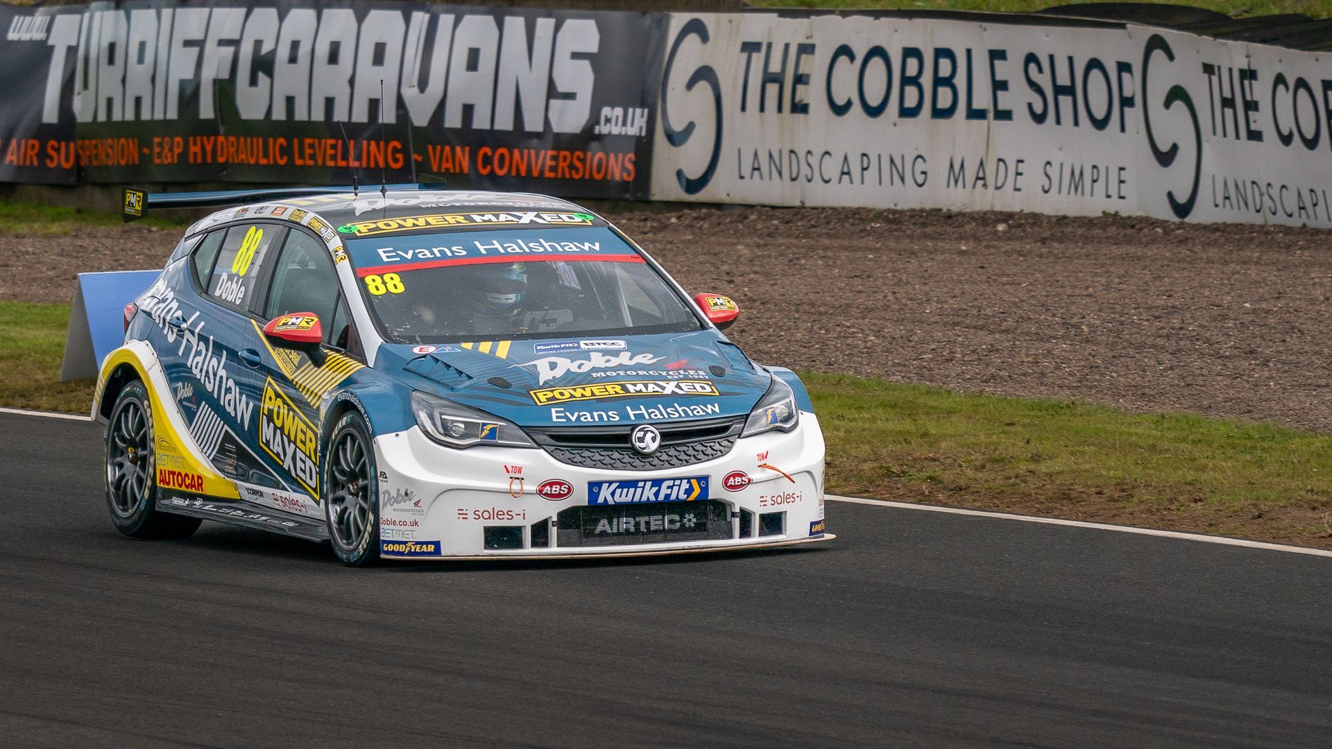 A race car is on a track in front of a sign that says the cobble shop