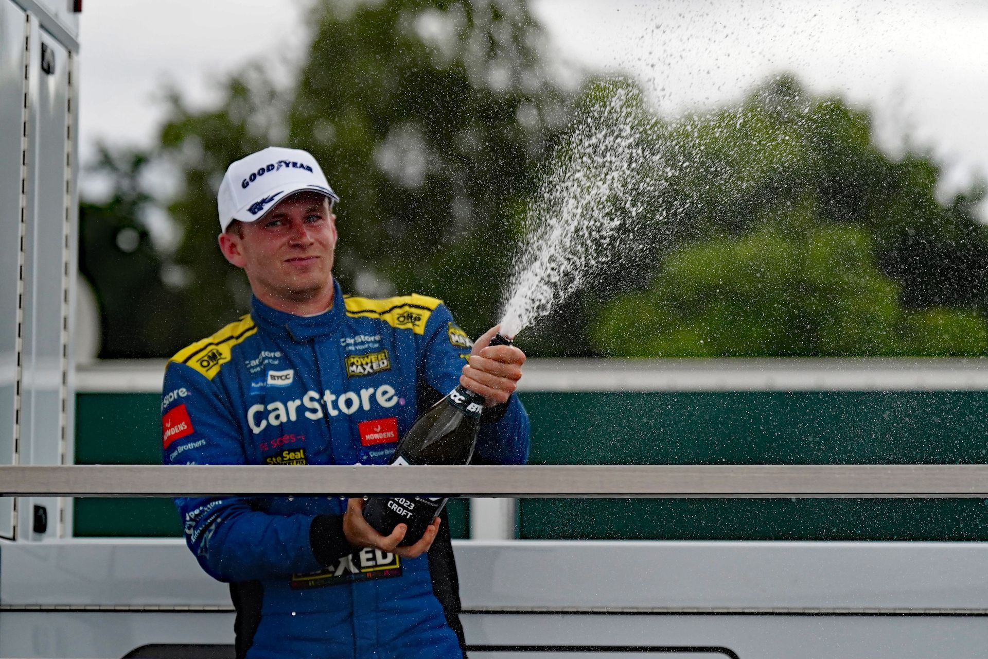 A man in a racing suit is holding a bottle of champagne and spraying it.