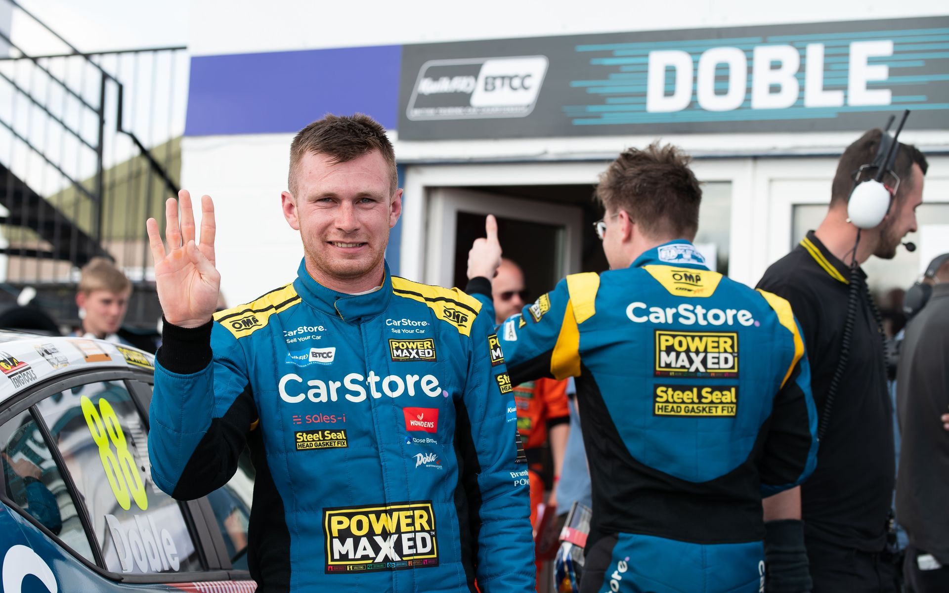 A man in a blue and yellow racing suit is standing in front of a car.