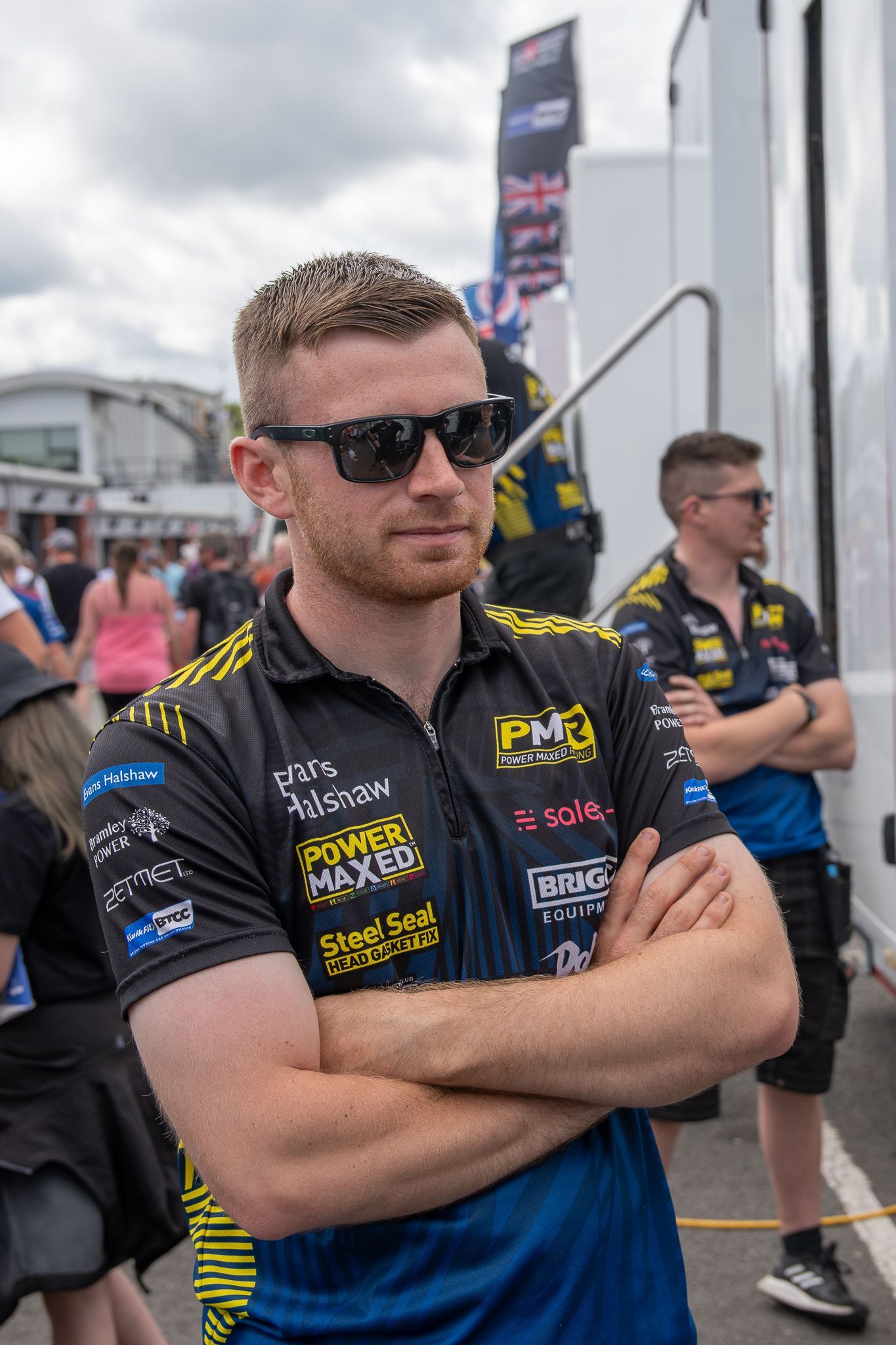 A man wearing sunglasses and a blue shirt is standing with his arms crossed.