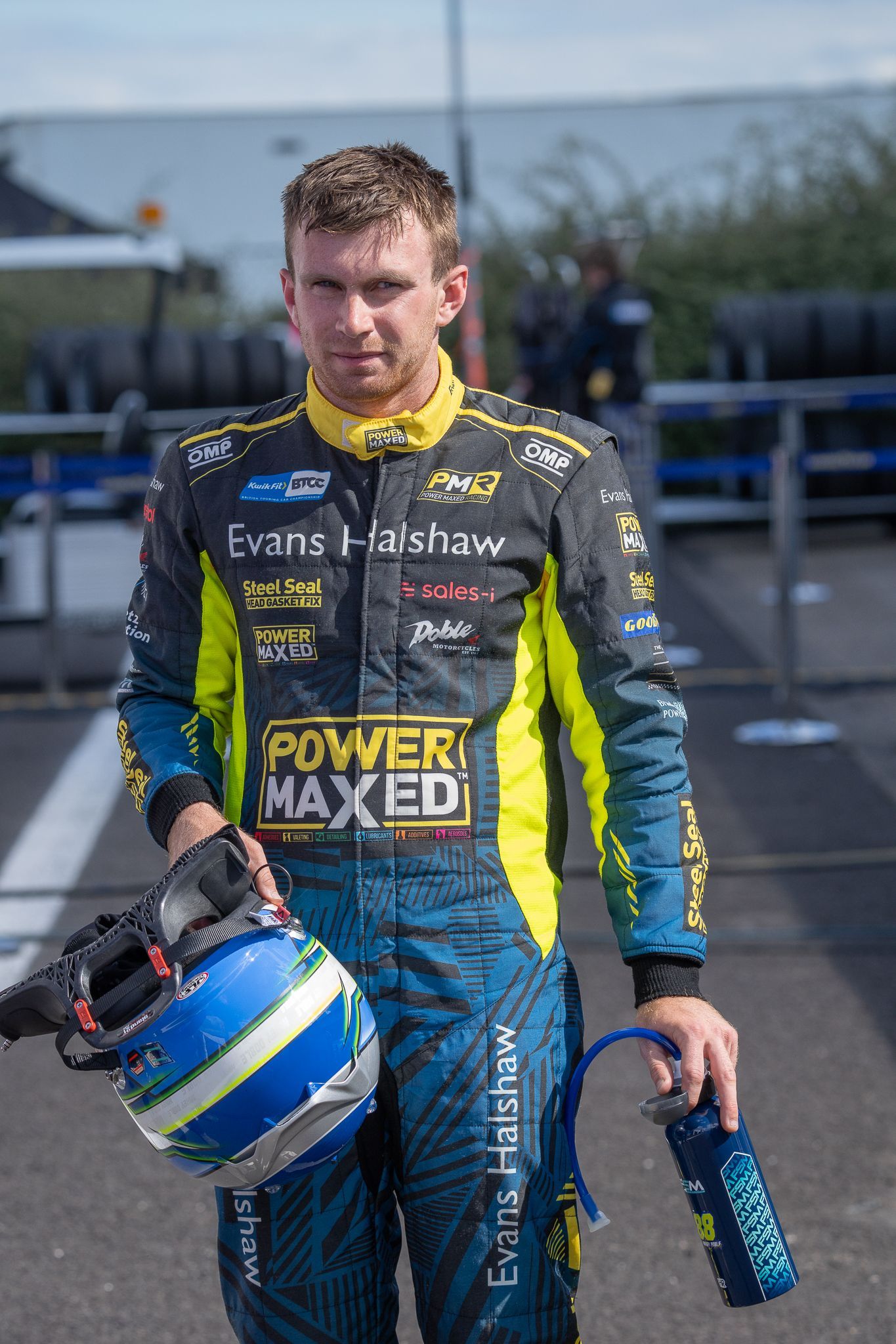 A man in a racing suit is holding a helmet and a water bottle.