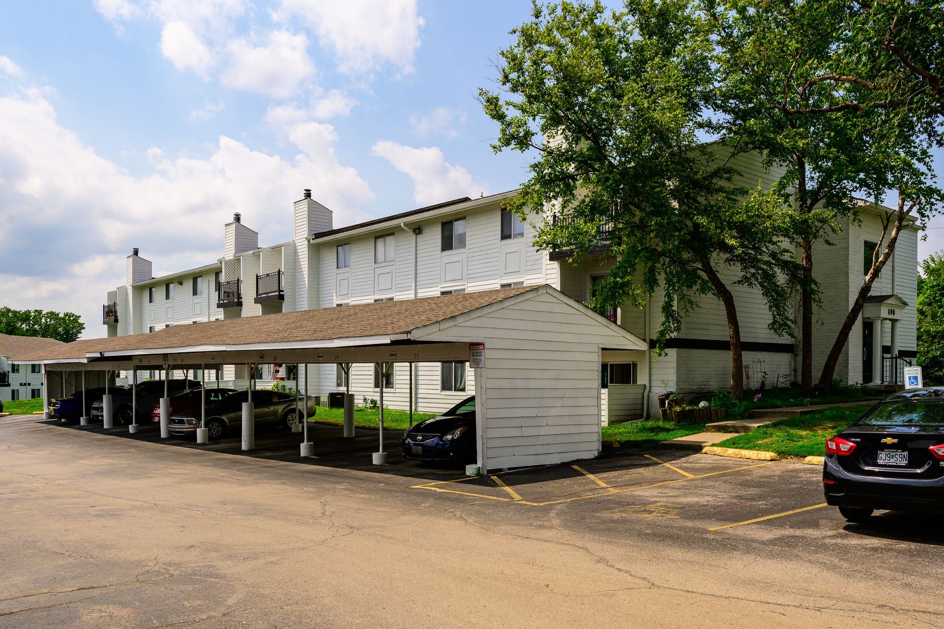 A row of cars are parked under a carport in front of a building.