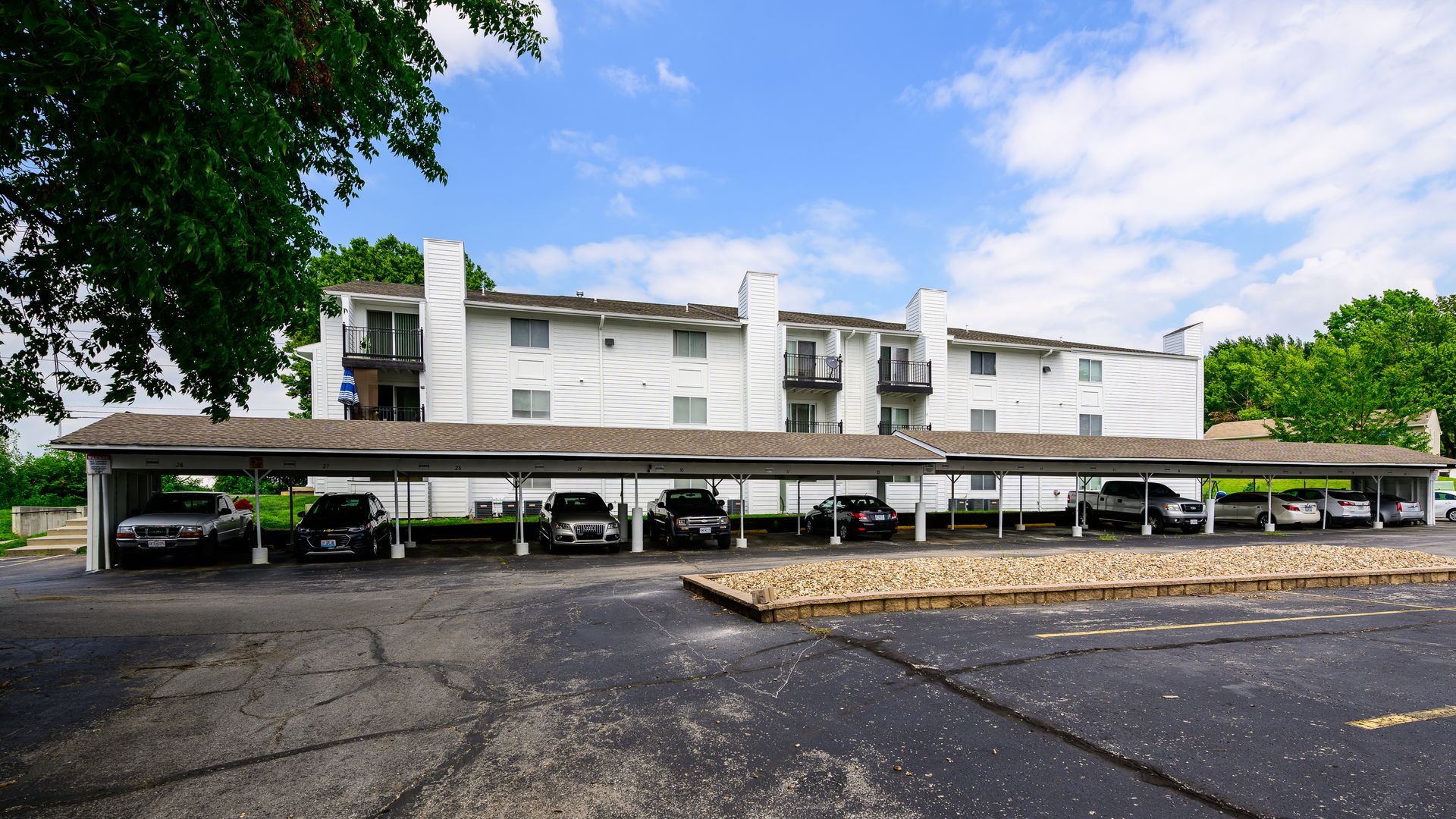 A row of cars are parked under a covered parking lot in front of a building.