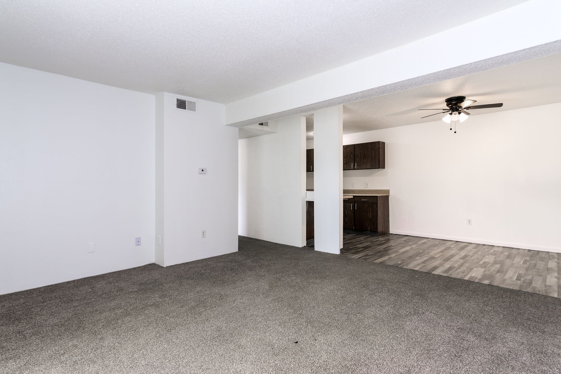 An empty living room with a ceiling fan and a kitchen in the background.