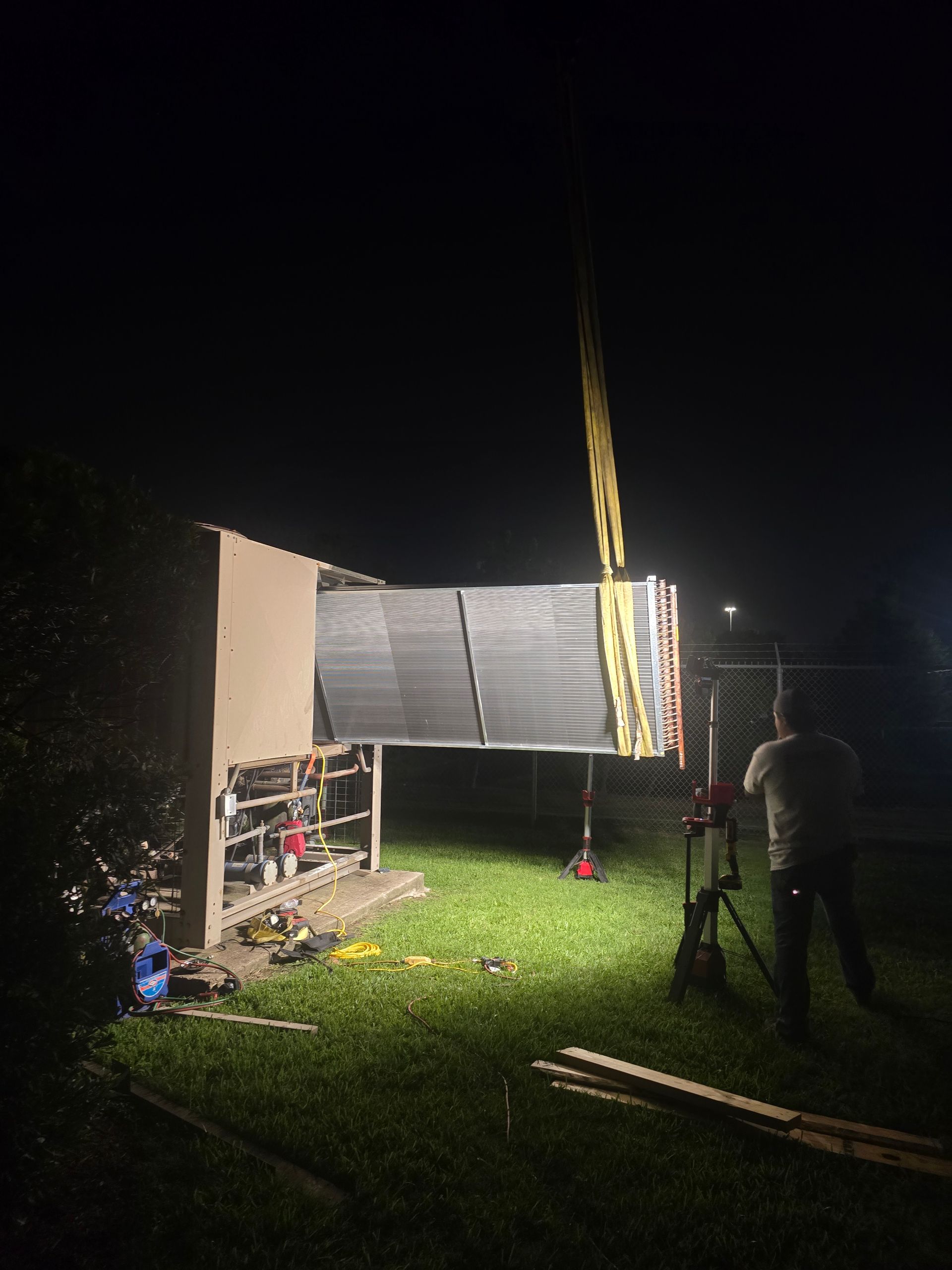 American Air Services, LLC - A large air conditioning unit being lifted by a crane at night; a worker stands nearby.