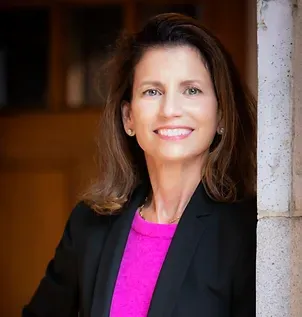 A woman in a pink sweater and black jacket is leaning against a wall and smiling.