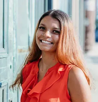 A woman in a red shirt is smiling and standing in front of a door.