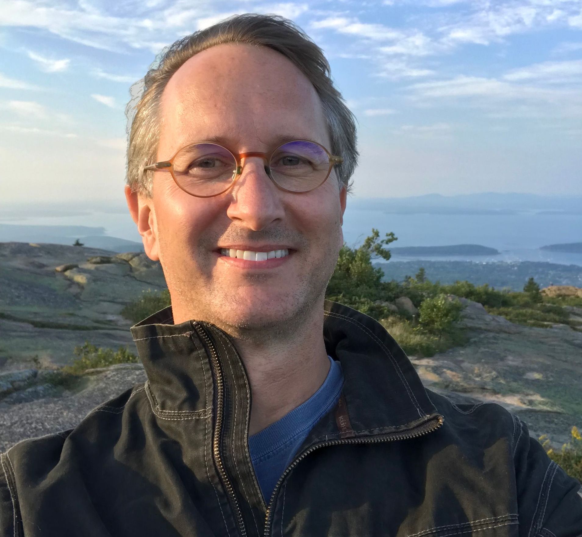 A man wearing glasses and a black jacket is smiling for the camera