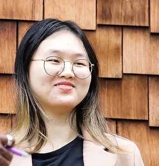 A woman wearing glasses and a jacket is holding a pen in front of a wooden wall.
