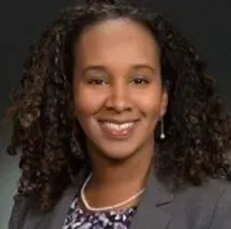 A woman with curly hair is wearing a gray jacket and a necklace.