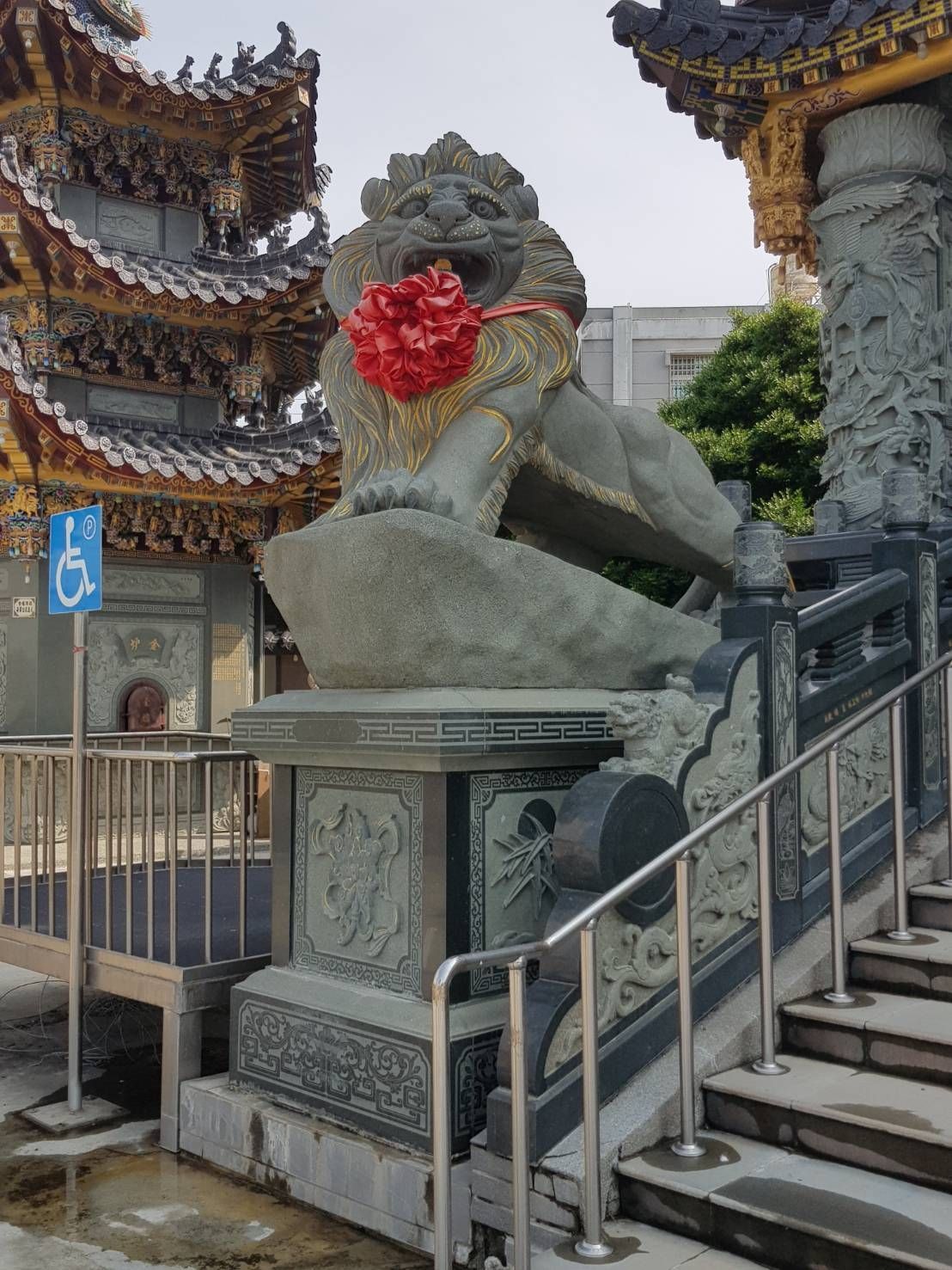 寺廟階梯旁,一座裝飾華麗的寶塔屋頂下,矗立著一尊飾有紅色花環的石獅雕像。